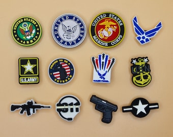 May include: A collection of twelve military and patriotic themed shoe charms. The set includes circular and shaped emblems featuring the United States Army, Navy, Marine Corps, Air Force, a veteran badge, a 911 emblem, a rifle, and a handgun.