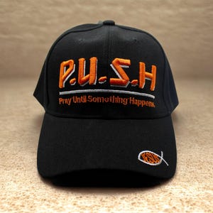 Op de afbeelding: Zwarte baseballcap met de geborduurde tekst "P.U.S.H. Pray Until Something Happens" in oranje en wit. Een klein oranje en wit vissymbool is op de zijkant geborduurd.