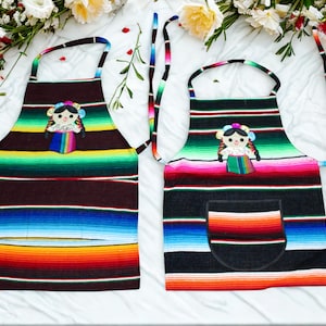 May include: Two colourful aprons with a striped serape design. Each apron features a doll applique and a pocket on one. The aprons have a rainbow of colours including red, orange, yellow, green, blue, and black.