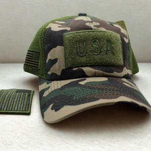 May include: A camouflage baseball cap with a green mesh back and a rectangular olive green patch on the front featuring the text USA in raised lettering. A small rectangular patch with a black and green American flag design sits to the left.