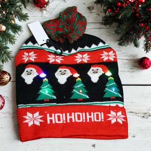 May include: A black, red, and white knitted Christmas beanie with a green and red pom-pom. The hat features Santa Claus faces and Christmas trees with lights. The text "HO! HO! HO!" is displayed on the front.
