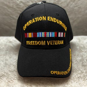 May include: Black baseball cap with embroidered text "OPERATION ENDURING FREEDOM VETERAN" in gold. Features a row of colorful ribbons above the text. The cap has a curved brim and a structured crown.