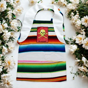 May include: A colorful apron with a striped design in shades of blue, green, red, and white. The apron features a white bib and straps. A rectangular patch with a skull and crossbones graphic and the text "LA CALAVERA" is centered on the apron.