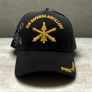 May include: A black baseball cap featuring yellow embroidered text that reads Air Defense Artillery above a crossed cannon and missile emblem. The brim displays the yellow embroidered text First To Fire, and a grey graphic is on the side.