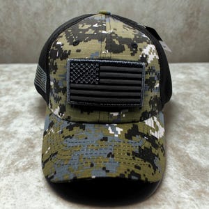 May include: A camouflage baseball cap with a black mesh back and a curved brim. The cap features a black and gray American flag patch on the front. The camouflage pattern includes shades of green, gray, and black.