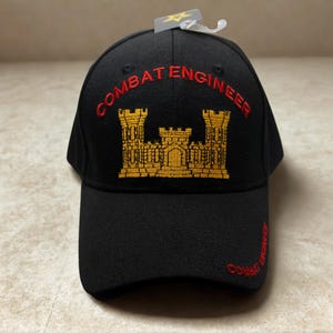 May include: Black baseball cap with "COMBAT ENGINEER" embroidered in red. The cap features a gold embroidered castle design and additional red text on the side. A tag is attached to the top.