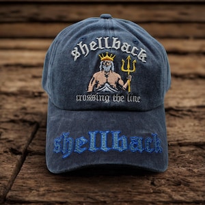 May include: A navy blue baseball cap with the word "shellback" embroidered in white script above an illustration of a muscular figure with a crown and trident. The phrase "crossing the line" is below the illustration, and "shellback" is embroidered in blue on the cap's brim.
