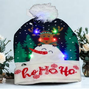 May include: A navy blue and white knit Christmas hat with a reindeer and snowman design. The hat features the text "ho ho ho" in red, and has a white pom-pom and LED lights.