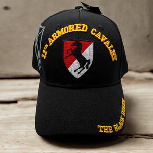 Puede incluir: Gorra de béisbol negra con texto bordado y escudo. La gorra presenta las palabras "11th ARMORED CAVALRY" en amarillo, arqueadas sobre un escudo con un caballo negro sobre un fondo rojo y blanco. También está bordado el texto "THE BLACK HORSE".