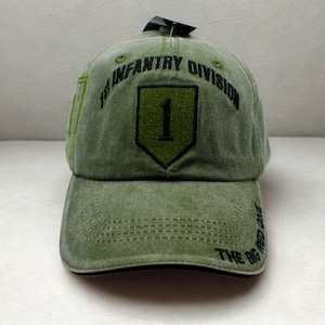 May include: Olive green baseball cap with the text "1st Infantry Division" and "The Big Red One". The cap features a shield with the number "1" in the center. The hat has a curved brim and a black underside.