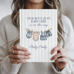 May include: A hardcover book with a light blue and white vertical striped cover. The text reads OUR BLUE JEAN BABY GIRL is on the way and Baby Emily. An illustration shows baby clothes hanging on a clothesline with wooden pins.