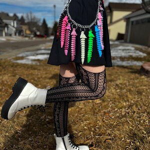 May include: A black dress with a silver chain belt adorned with colorful, segmented ornaments. The outfit is paired with white platform boots and patterned stockings. The background shows a residential area with a blue sky.