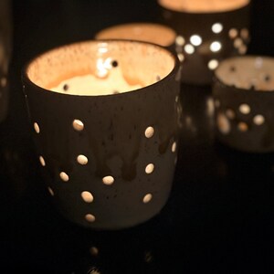 May include: A set of cylindrical ceramic candle holders with a speckled off-white glaze and a series of circular cutouts. Each holder features a warm, glowing light emanating from within, casting a soft illumination through the perforated holes.