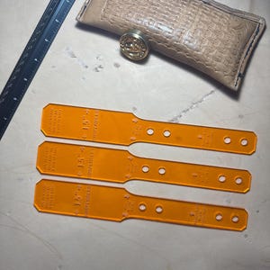 May include: An assortment of leatherworking tools, including three orange 15-inch acrylic rulers, a black ruler, and a tan leather case with a decorative clasp. The tools are arranged on a light brown surface, suggesting a craft or workshop setting.