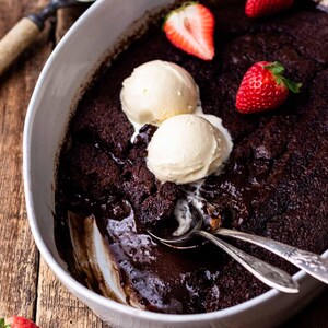 May include: A white oval dish of chocolate cake, garnished with vanilla ice cream and strawberries. Two spoons are in the cake. The dish is on a wooden surface. A dessert.
