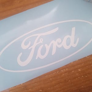 May include: A white Ford logo decal on a light blue background. The Ford logo is in a classic script font, enclosed within an oval. The decal is on a wooden surface.