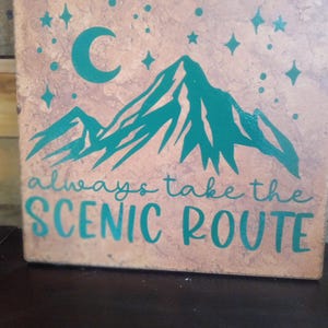 May include: A square, decorative tile with a teal mountain range, crescent moon, and stars design. The phrase "always take the SCENIC ROUTE" is written in teal script. The tile has a textured, mottled, tan and brown surface.