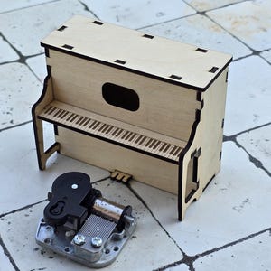 Może przedstawiać: Miniaturowy drewniany fortepian z wycinanym laserowo wzorem, z klawiaturą i oddzielną mechaniczną pozytywką. Fortepian wykonany jest z jasnego drewna i ma prostokątny kształt. Pozytywka jest srebrna i czarna.