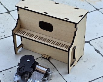 May include: A miniature wooden piano with a laser-cut design, featuring a keyboard and a separate mechanical music box. The piano is made of light-colored wood and has a rectangular shape. The music box is silver and black.