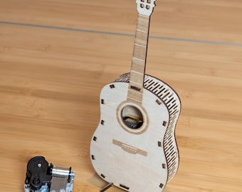 May include: A wooden guitar music box with a detailed design, featuring a sound hole and fretboard. The guitar stands upright on a wooden surface, next to a separate mechanical music box component. The guitar is made of light-colored wood.