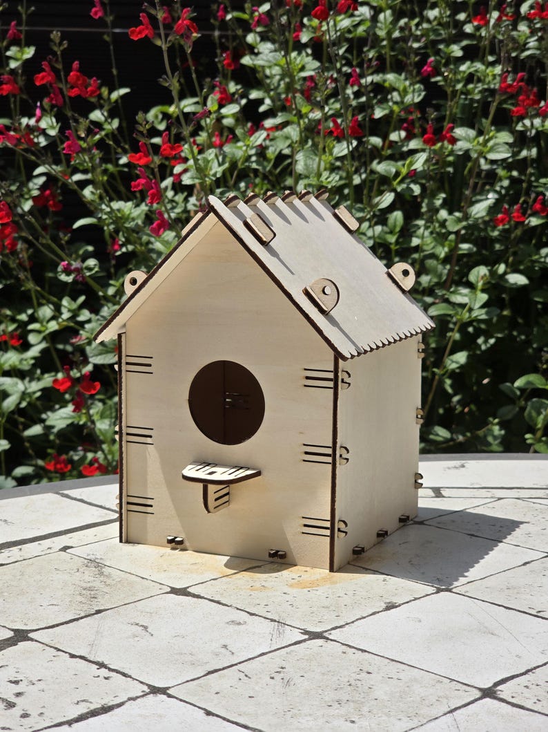 May include: A wooden birdhouse with a natural finish, featuring a round entry hole and a small perch. The birdhouse has a gabled roof and is constructed with visible joinery details. The background includes red flowers and green foliage.