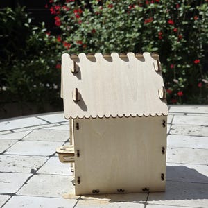 May include: A small, unfinished wooden birdhouse with a scalloped roof and small, rectangular side pieces. The birdhouse is assembled with small metal hinges and is sitting on a stone surface. The background features green foliage and red flowers.