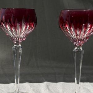 May include: Two wine glasses featuring deep red bowls with vertical cut glass detailing that transitions into clear, faceted stems and circular bases. The design showcases a contrast between the colored upper section and the transparent lower stem.