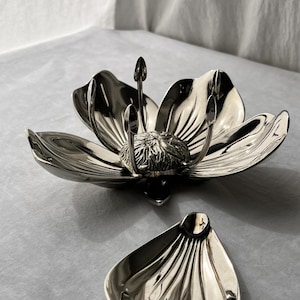 May include: A polished silver-toned metal bowl shaped like an open flower with six large petals and a textured central stamen, accompanied by a separate, smaller matching petal-shaped dish with a smooth, reflective surface and ribbed detailing.