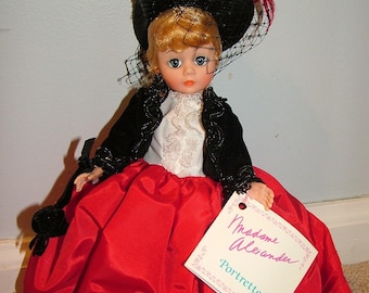 May include: A red and black Madame Alexander doll wearing a black velvet jacket, a white lace blouse, and a red satin skirt. The doll is wearing a black hat with a veil and a red feather. The doll is holding a tag that says "Madame Alexander Portraittes".