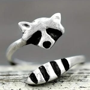 May include: A silver ring with a raccoon design. The raccoon's face is on the top of the ring, and its tail wraps around the band.