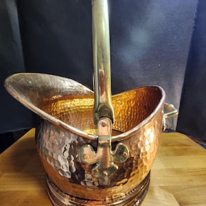 May include: A hammered copper coal scuttle with a polished brass handle and a fleur-de-lis attachment. The vessel features a wide, open top, a circular base, and a metallic finish that reflects light across its textured, dimpled surface.
