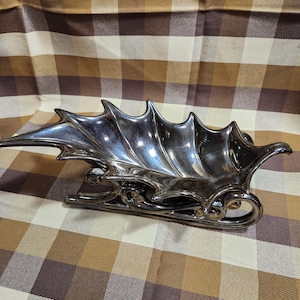 May include: A silver decorative sleigh with a leaf-like design on the sides. The sleigh has a curved runner design and is placed on a plaid patterned surface.