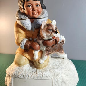 Peut inclure: Figurine en céramique représentant une personne en parka jaune avec une capuche bordée de fourrure blanche, tenant un chien marron et blanc. La figurine est posée sur une base enneigée avec un espace rectangulaire vierge.