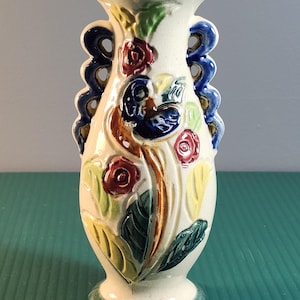 May include: Cream ceramic vase with a flared top and pedestal base. The vase is decorated with colourful floral and leaf designs in red, blue, yellow, and green. It has two blue, scalloped handles on either side.