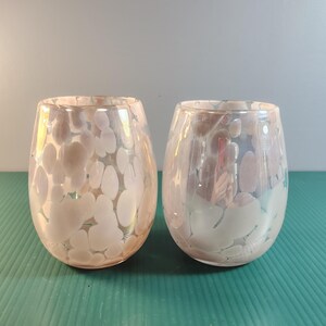 May include: Two stemless wine glasses with an iridescent finish and a pattern of white, irregular shapes. The glasses have a rounded shape and are made of clear glass. The background is a solid light blue.