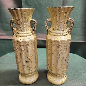 May include: Two gold-coloured vases with a textured, droplet-like pattern. Each vase has a flared top, two small handles, and a pedestal base. The vases are displayed on a green surface.