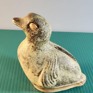 May include: A ceramic duck figurine in shades of beige and brown. The duck has a textured surface, a detailed beak, and a slot on its back. The figurine is a decorative item, possibly a bank or a collectible.
