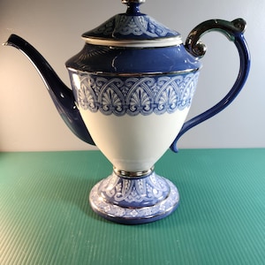 May include: A blue and white porcelain teapot with a decorative design. The teapot has a curved spout, a handle with a silver accent, and a lid with a gold finial. The base of the teapot is also decorated with a matching design.