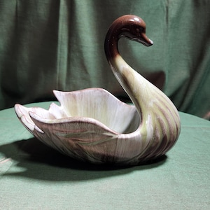 May include: A ceramic swan-shaped bowl featuring a glossy finish with a gradient color scheme. The neck and head are a deep brown, transitioning into a lighter cream and sage green body with molded feather details along the wings and base.