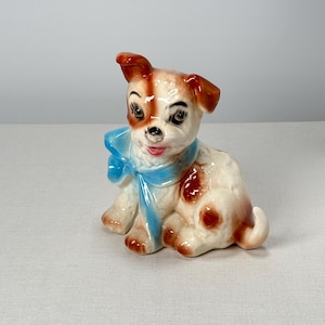 May include: A ceramic figurine of a brown and white puppy wearing a blue bow. The puppy is sitting with its head tilted and has a happy expression.