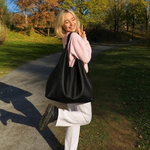 Black oversized leather tote bag styled on a smiling woman outdoors during golden hour autumn scenery. Minimalist slouchy shoulder bag with soft genuine leather texture, large capacity everyday purse, quiet luxury fashion aesthetic, modern hobo tote for women, chic street style handbag, casual fall outfit inspiration, trendy oversized carryall bag, elegant soft leather shoulder purse, commuter tote, laptop bag, designer inspired fashion accessory, neutral capsule wardrobe essential