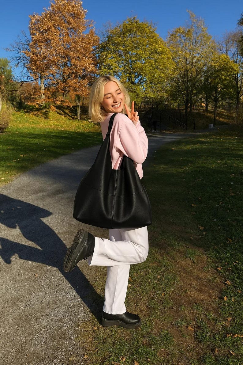 Black oversized leather tote bag styled on a smiling woman outdoors during golden hour autumn scenery. Minimalist slouchy shoulder bag with soft genuine leather texture, large capacity everyday purse, quiet luxury fashion aesthetic, modern hobo tote for women, chic street style handbag, casual fall outfit inspiration, trendy oversized carryall bag, elegant soft leather shoulder purse, commuter tote, laptop bag, designer inspired fashion accessory, neutral capsule wardrobe essential