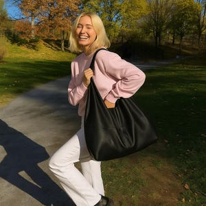 Woman carrying an oversized black leather hobo tote bag outdoors during golden hour in a beautiful autumn park setting. Soft slouchy shoulder bag with minimalist design, genuine leather texture, large capacity everyday purse, quiet luxury handbag aesthetic, trendy oversized tote for women, chic commuter bag, modern street style fashion, casual fall outfit inspiration, designer inspired leather shoulder purse, elegant carryall handbag, stylish work tote, laptop bag, handmade leather hobo bag
