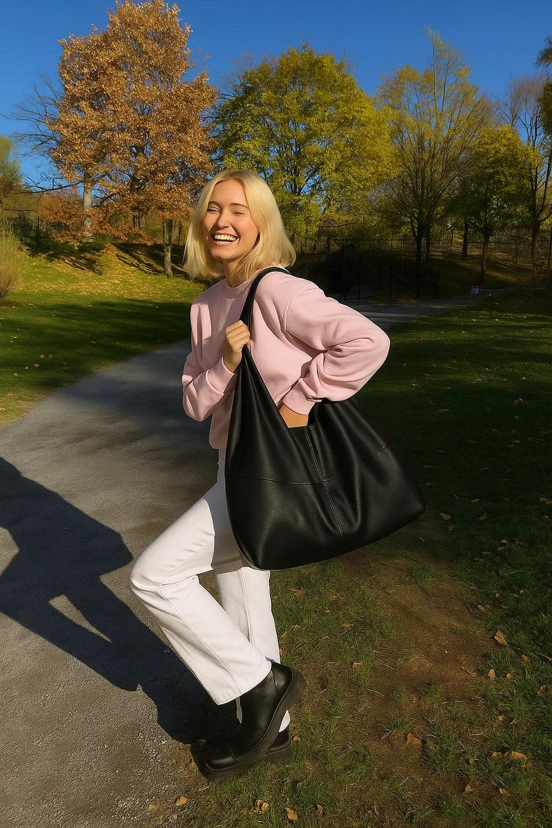 Woman carrying an oversized black leather hobo tote bag outdoors during golden hour in a beautiful autumn park setting. Soft slouchy shoulder bag with minimalist design, genuine leather texture, large capacity everyday purse, quiet luxury handbag aesthetic, trendy oversized tote for women, chic commuter bag, modern street style fashion, casual fall outfit inspiration, designer inspired leather shoulder purse, elegant carryall handbag, stylish work tote, laptop bag, handmade leather hobo bag