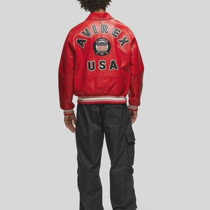 Back view of men’s red vintage leather bomber jacket showcasing large Avirex USA embroidered patch artwork across the back panel. Crafted from high quality genuine sheepskin leather, this retro aviator jacket features ribbed cuffs, ribbed waistband, and relaxed oversized streetwear fit. The bold red leather color gives a standout urban fashion appearance suitable for casual outfits, streetwear looks, and winter fashion styling.