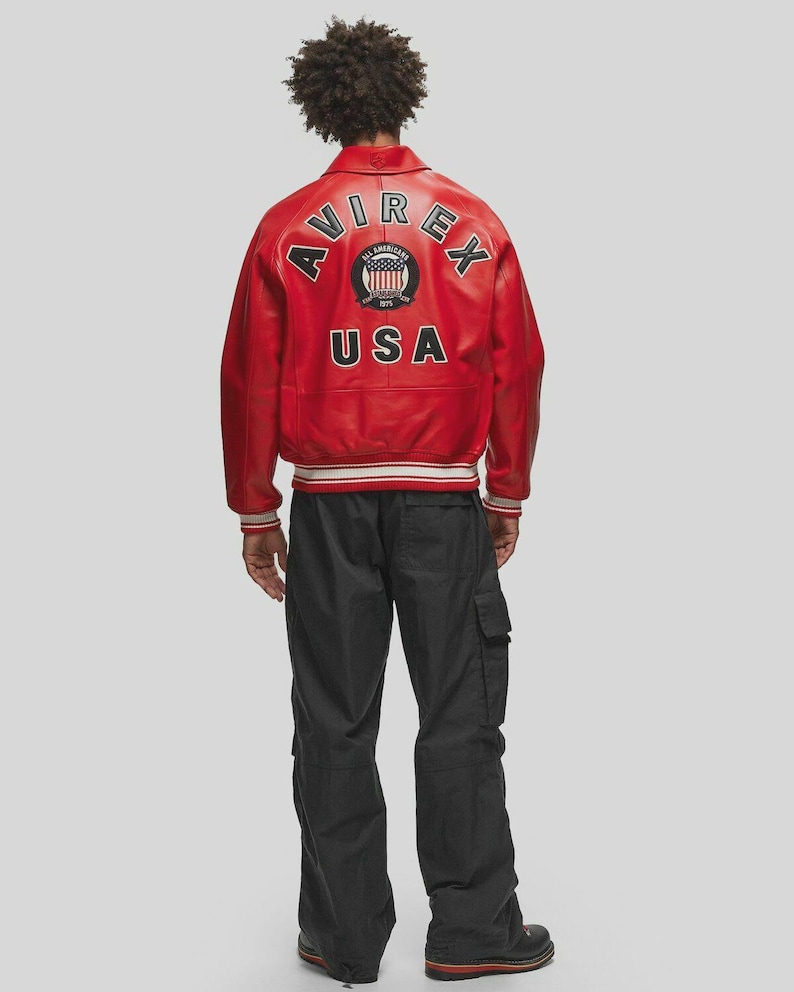 Back view of men’s red vintage leather bomber jacket showcasing large Avirex USA embroidered patch artwork across the back panel. Crafted from high quality genuine sheepskin leather, this retro aviator jacket features ribbed cuffs, ribbed waistband, and relaxed oversized streetwear fit. The bold red leather color gives a standout urban fashion appearance suitable for casual outfits, streetwear looks, and winter fashion styling.