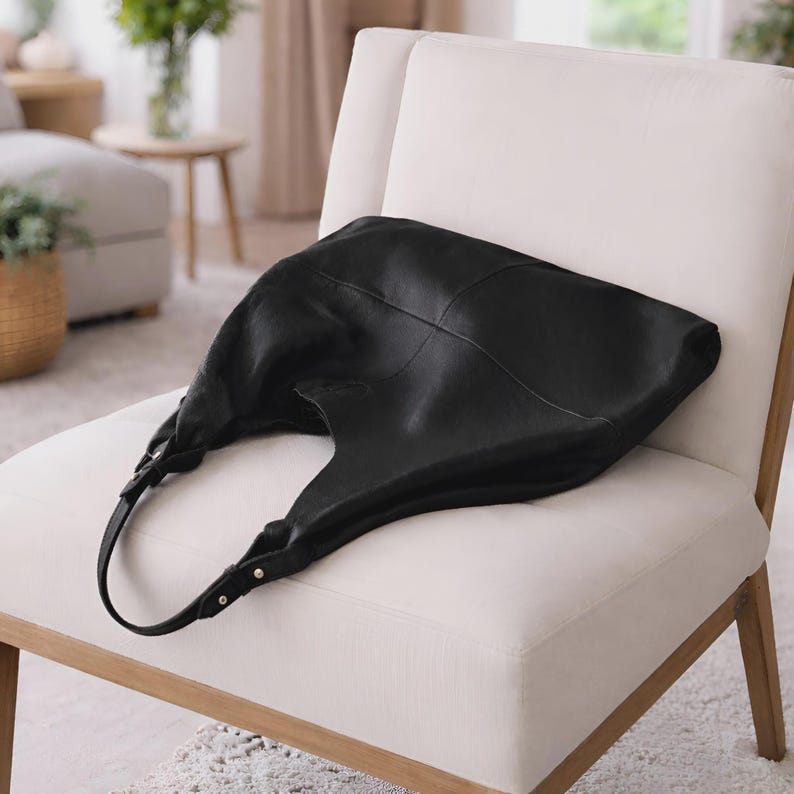 Oversized black leather hobo bag displayed on a modern cream accent chair in a minimalist luxury interior. Soft slouchy shoulder tote featuring genuine leather texture, spacious large capacity design, adjustable straps, and elegant quiet luxury aesthetic. Chic everyday handbag for women perfect for work, travel, shopping, and casual wear. Stylish oversized leather shoulder bag with modern minimalist fashion appeal, designer inspired carryall tote purse, handmade leather handbag, laptop friendly