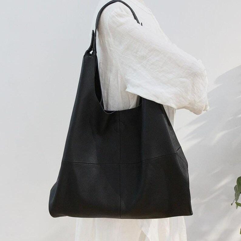 Minimalist black leather hobo tote bag with oversized slouchy silhouette displayed against a soft neutral background. Elegant genuine leather shoulder bag featuring a soft pebbled texture, spacious large capacity interior, modern quiet luxury aesthetic, chic everyday handbag for women, stylish carryall tote purse, designer inspired leather bag, casual minimalist fashion accessory, trendy oversized shoulder tote, laptop friendly work bag, handmade leather purse, capsule wardrobe essential