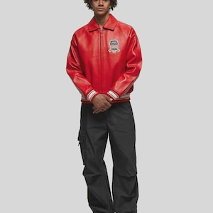 Men’s red genuine sheepskin leather bomber jacket front view featuring bold vintage aviator inspired design with embroidered chest patches and classic zip-up closure. The oversized streetwear fit combined with ribbed cuffs and waistband creates a timeless bomber silhouette. This premium red leather jacket is perfect for urban street fashion, casual outfits, fall and winter wear, and modern men’s outerwear styling.