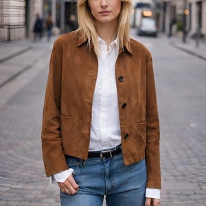 May include: A brown suede jacket with a collared neckline and button closure, worn over a white shirt. The outfit includes blue jeans and a black belt. The background shows a city street.